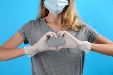 Woman in medical gloves and protective mask making heart with hands on light blue background, closeup