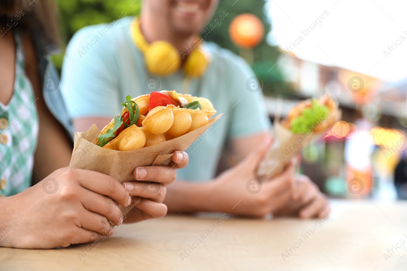 Photo of Young couple holding delicious bubble waffles with tomato and arugula at table outdoors, closeup