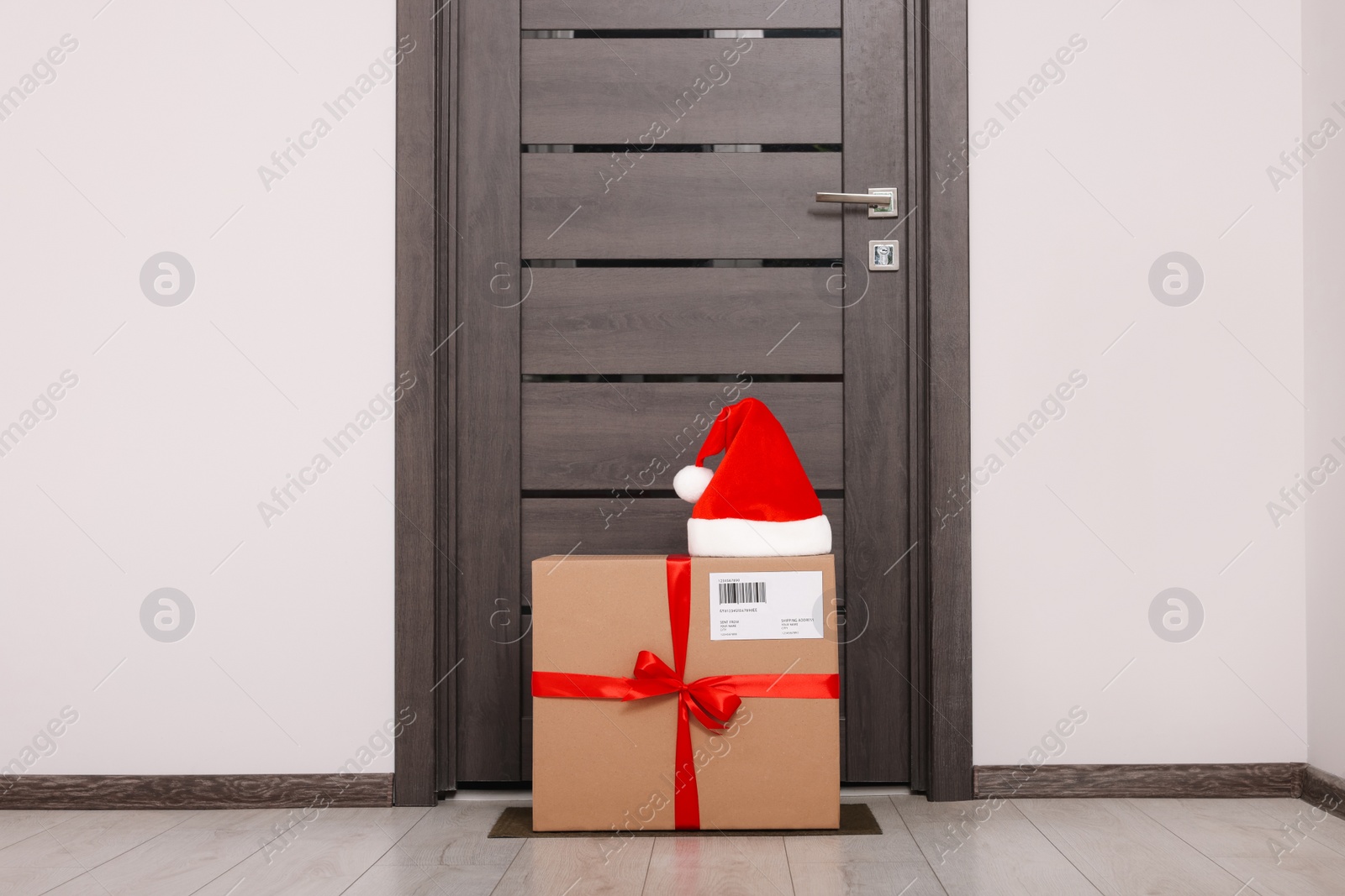 Photo of Christmas gift box and Santa hat near door indoors. Sending present by mail