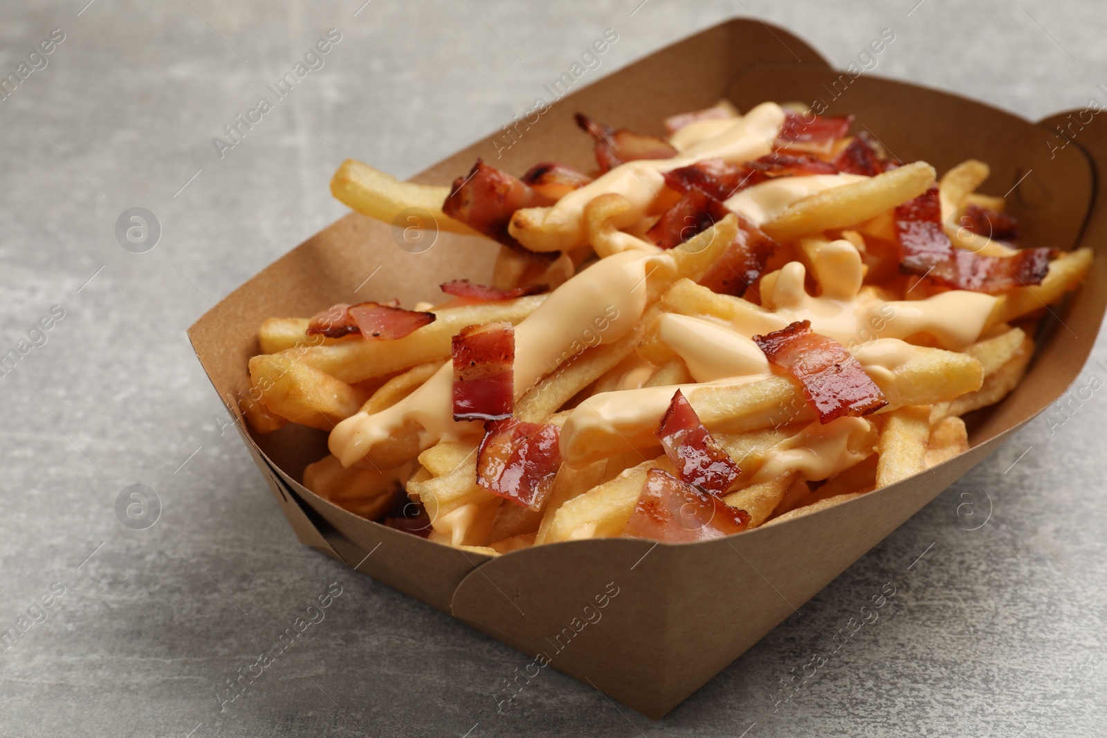 Photo of Tasty potato fries, cheese sauce and bacon in paper container on grey table, closeup