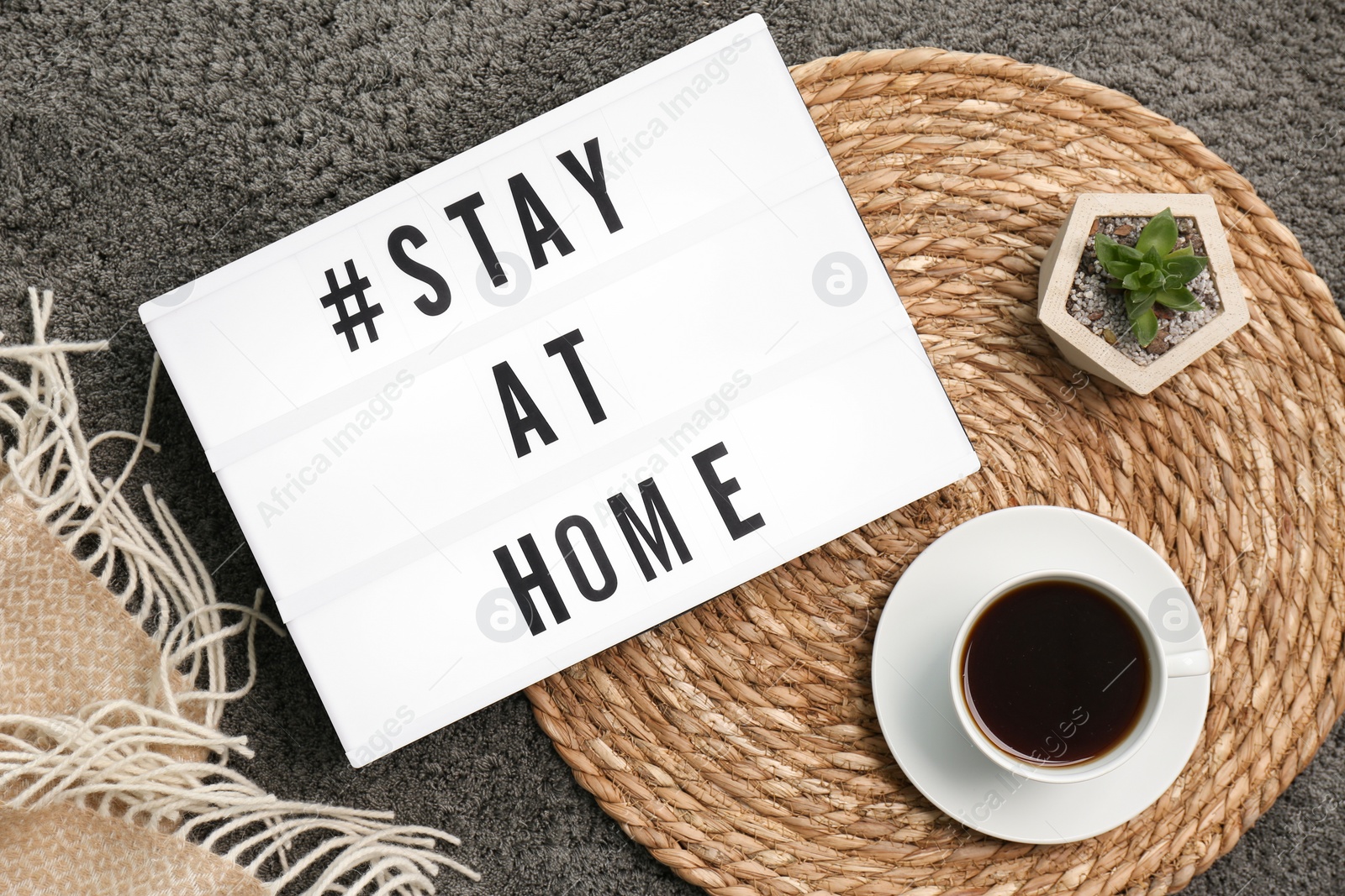 Photo of Cup of coffee, houseplant and lightbox with hashtag STAY AT HOME on grey carpet, flat lay. Message to promote self-isolation during COVID‑19 pandemic