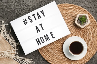 Photo of Cup of coffee, houseplant and lightbox with hashtag STAY AT HOME on grey carpet, flat lay. Message to promote self-isolation during COVID‑19 pandemic