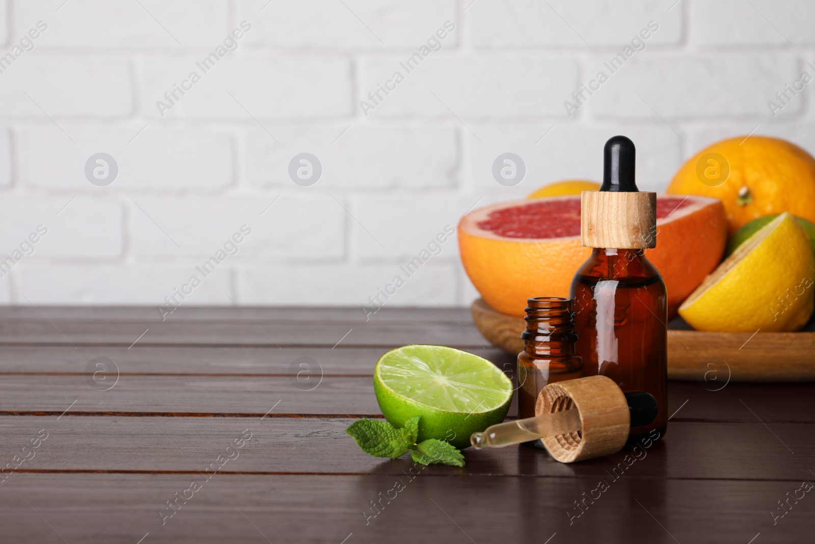 Photo of Bottles of essential oils with different citrus fruits and mint on wooden table against white brick wall. Space for text