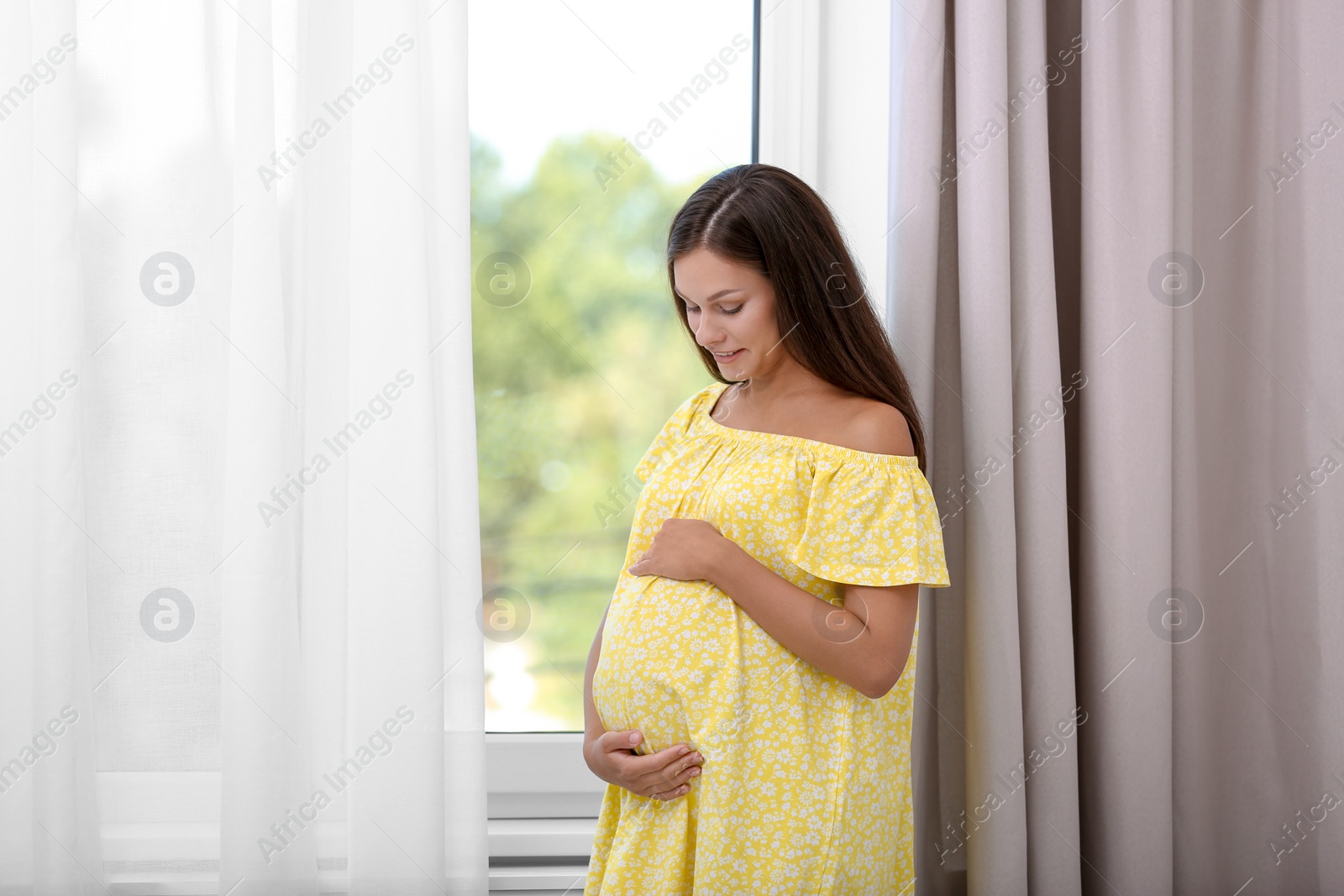 Photo of Happy pregnant woman standing near window at home