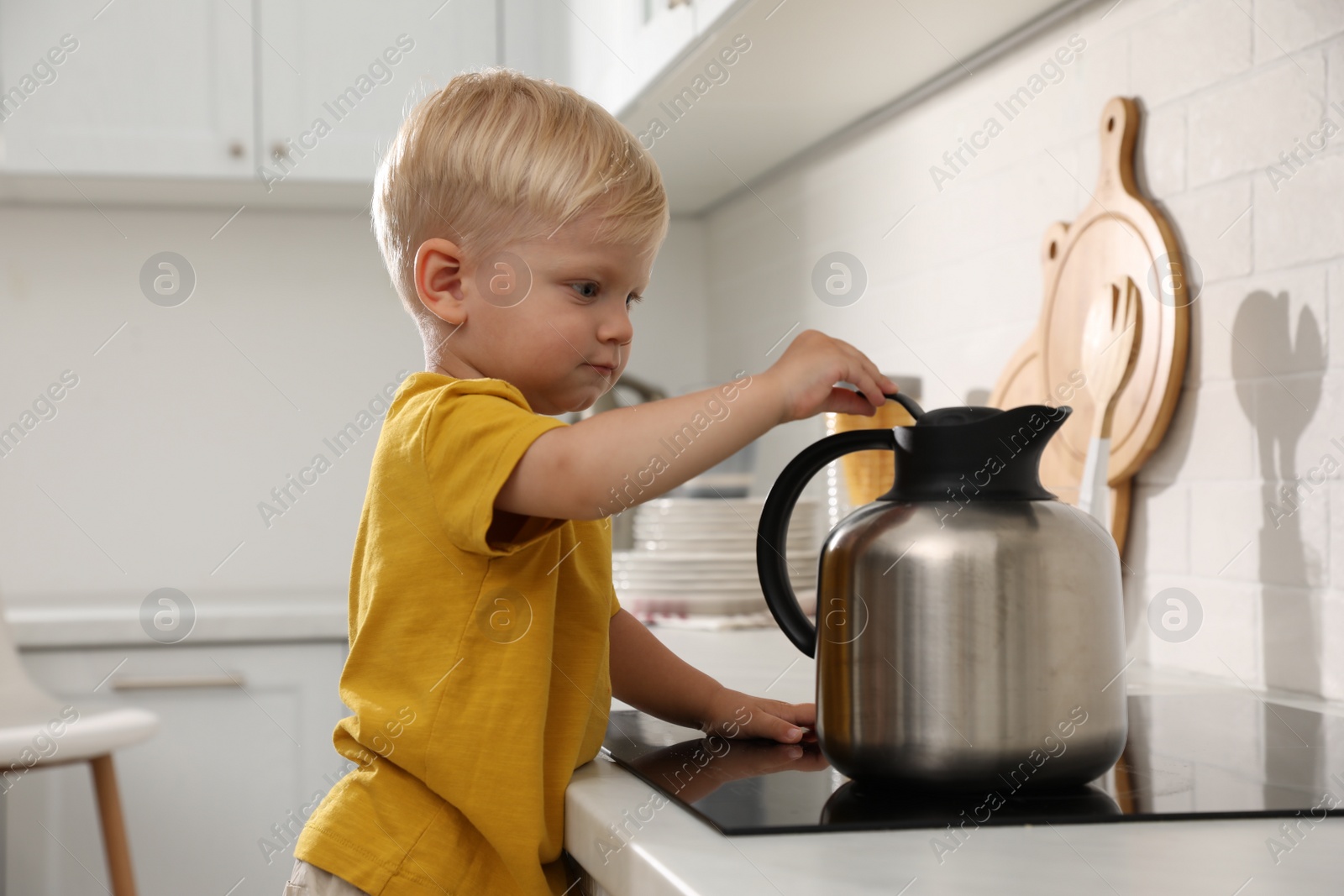 Photo of Curious little boy playing with kettle on electric stove in kitchen