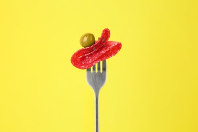 Fork with tasty slice of salami and olive on yellow background