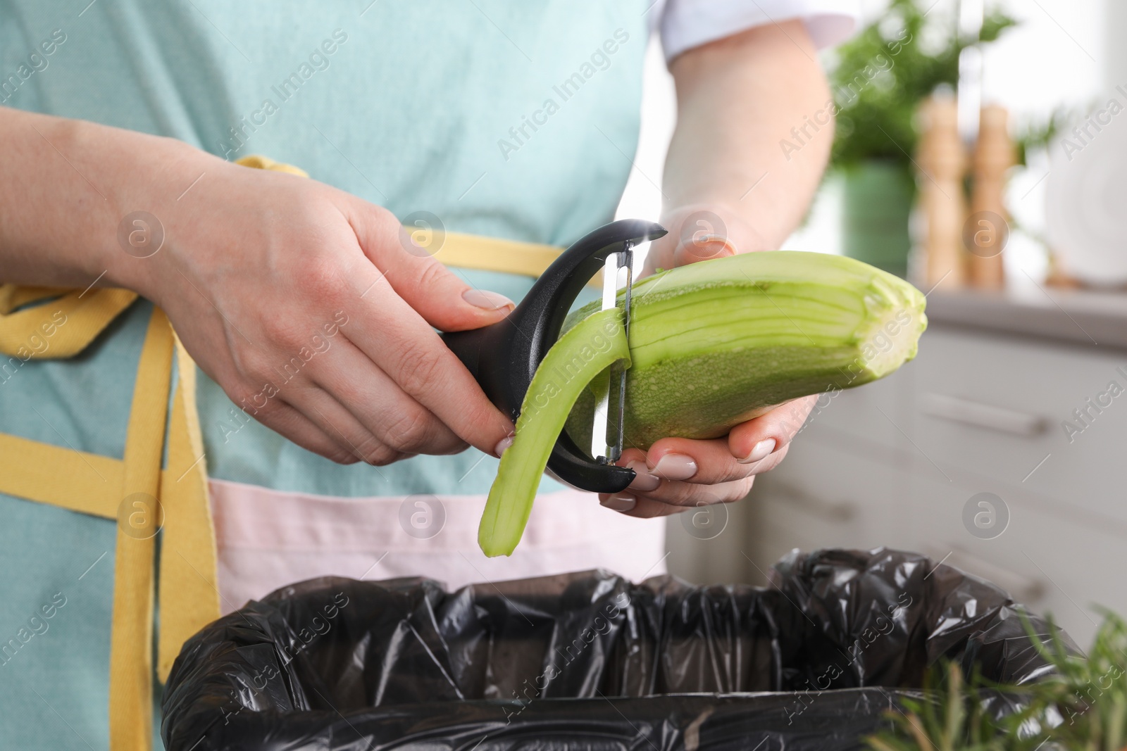 Photo of Woman peeling fresh zucchini above garbage bin indoors, closeup