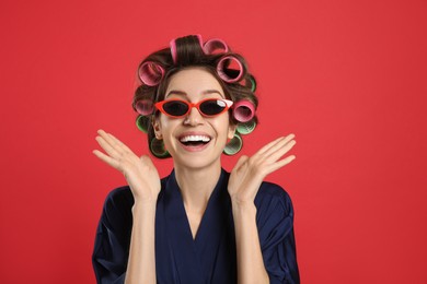 Beautiful young woman in silk bathrobe with hair curlers and sunglasses on red background