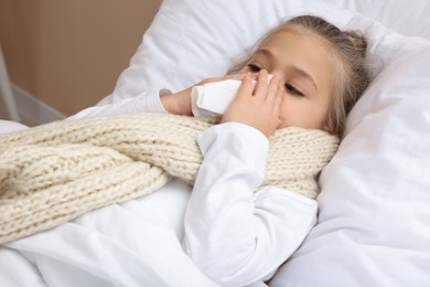 Photo of Sick girl with scarf and tissue lying in bed while blowing nose indoors, closeup