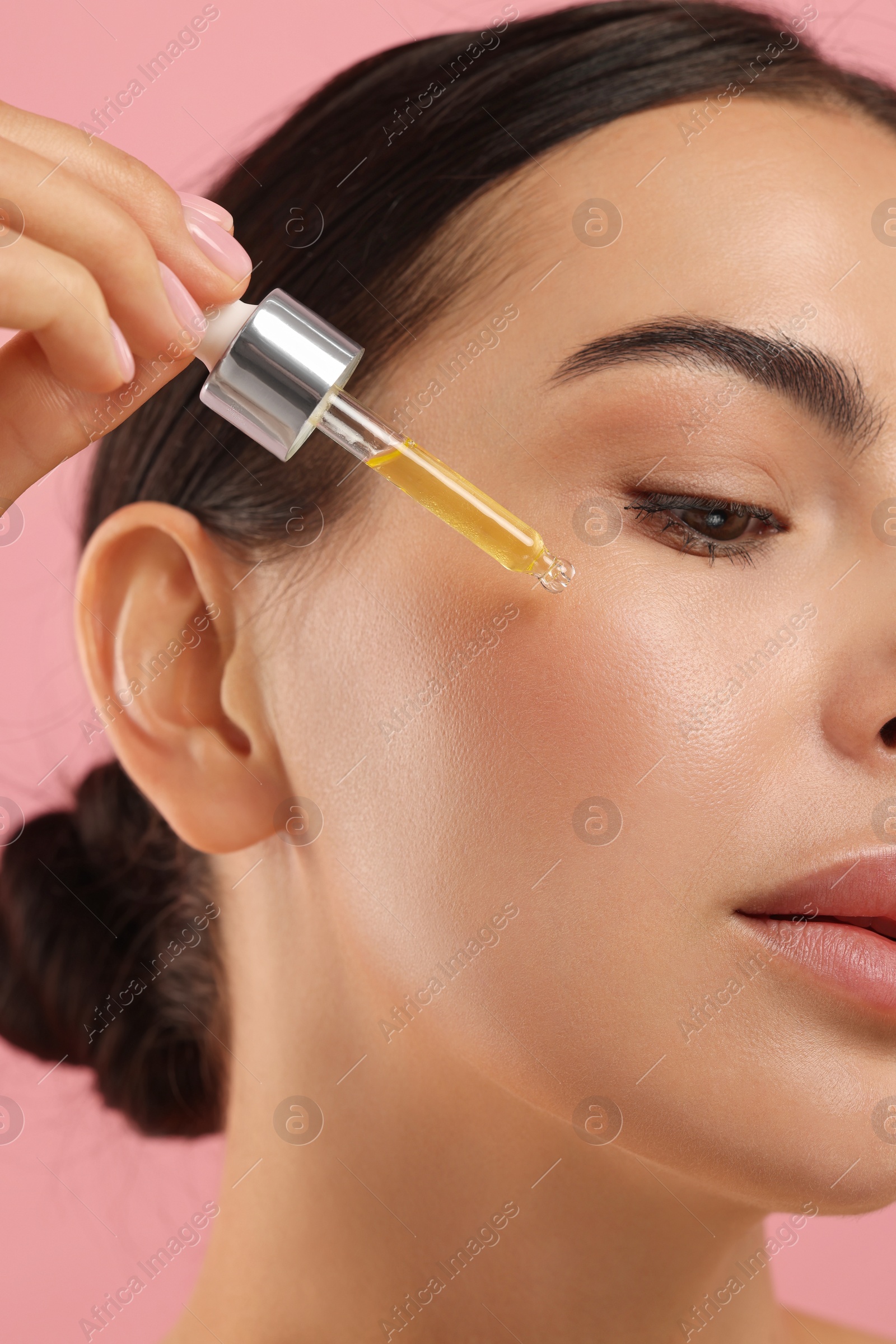 Photo of Beautiful young woman applying serum onto her face on pink background, closeup