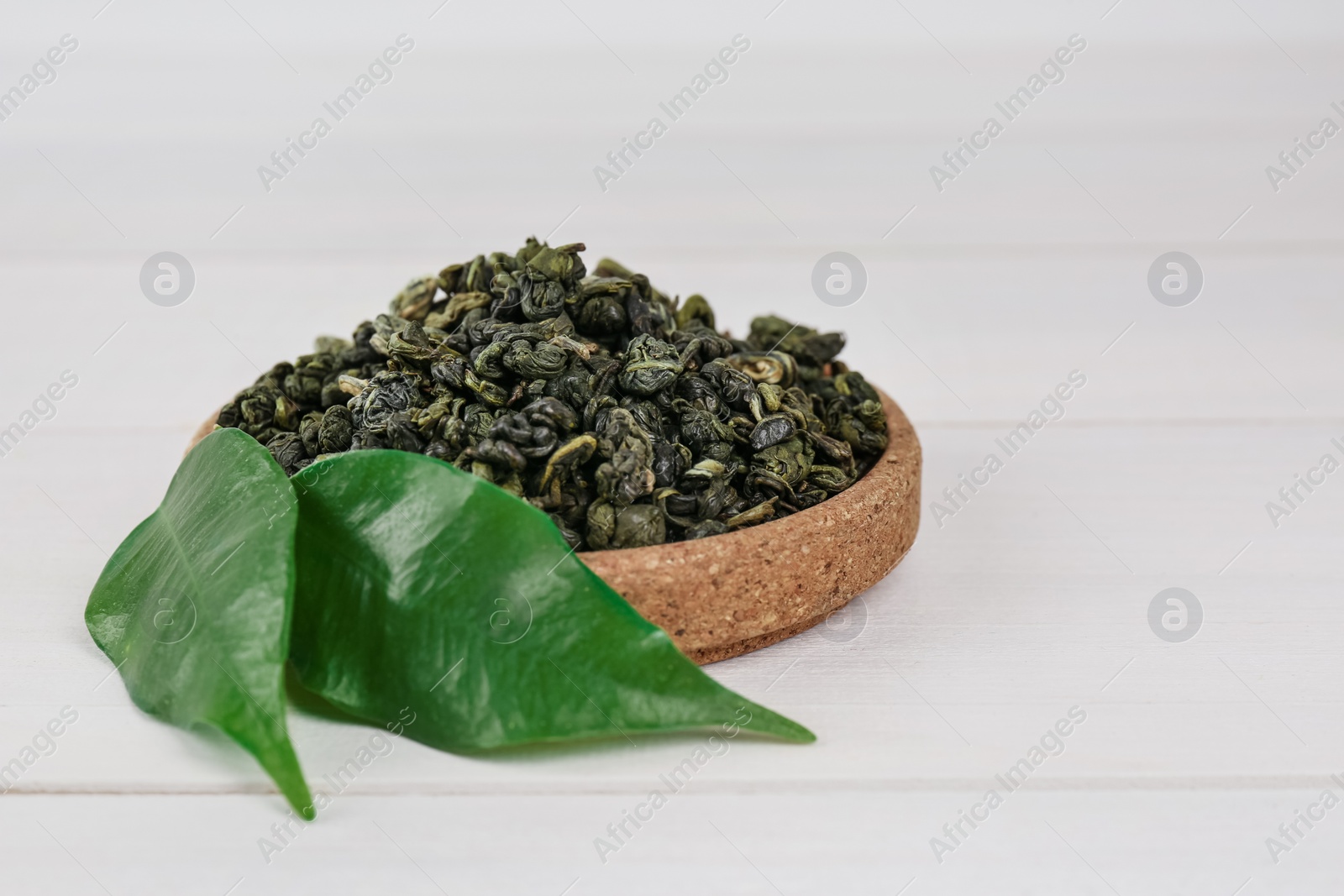 Photo of Dried green tea leaves in cork bowl on white wooden table