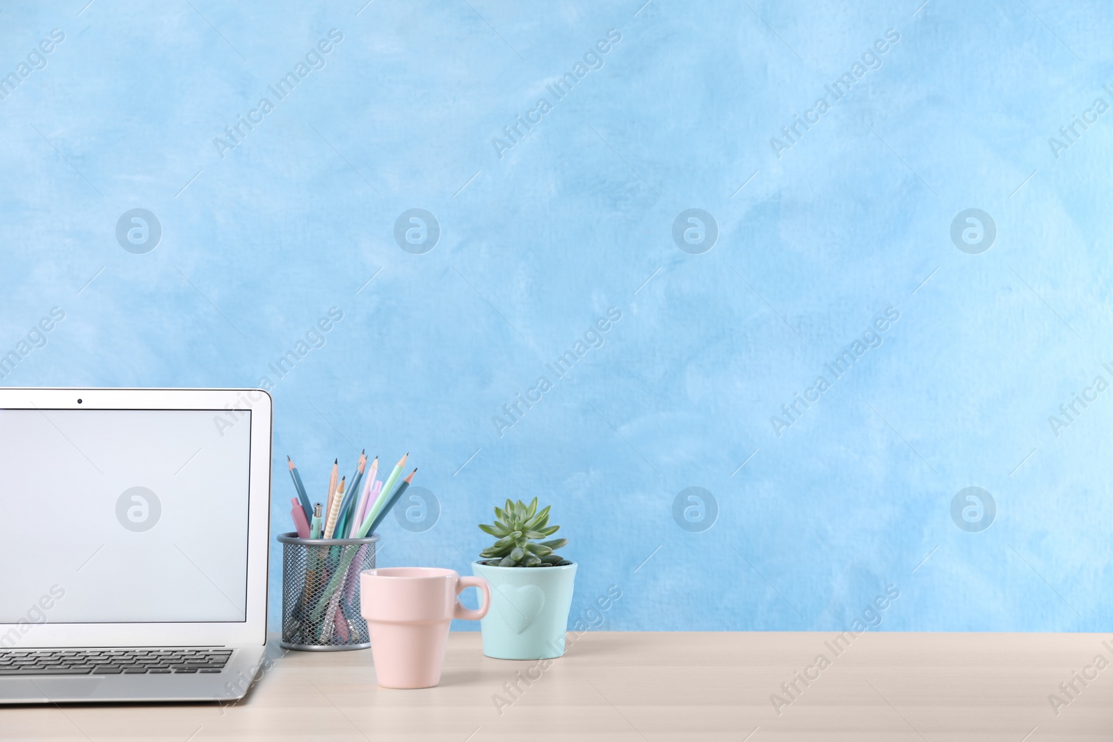 Photo of Modern laptop with blank screen, cup and office supplies on white table near light blue wall, space for text