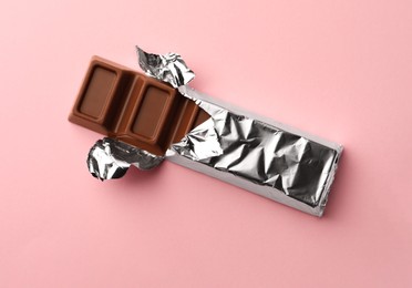 Delicious chocolate bar wrapped in foil on pink background, top view