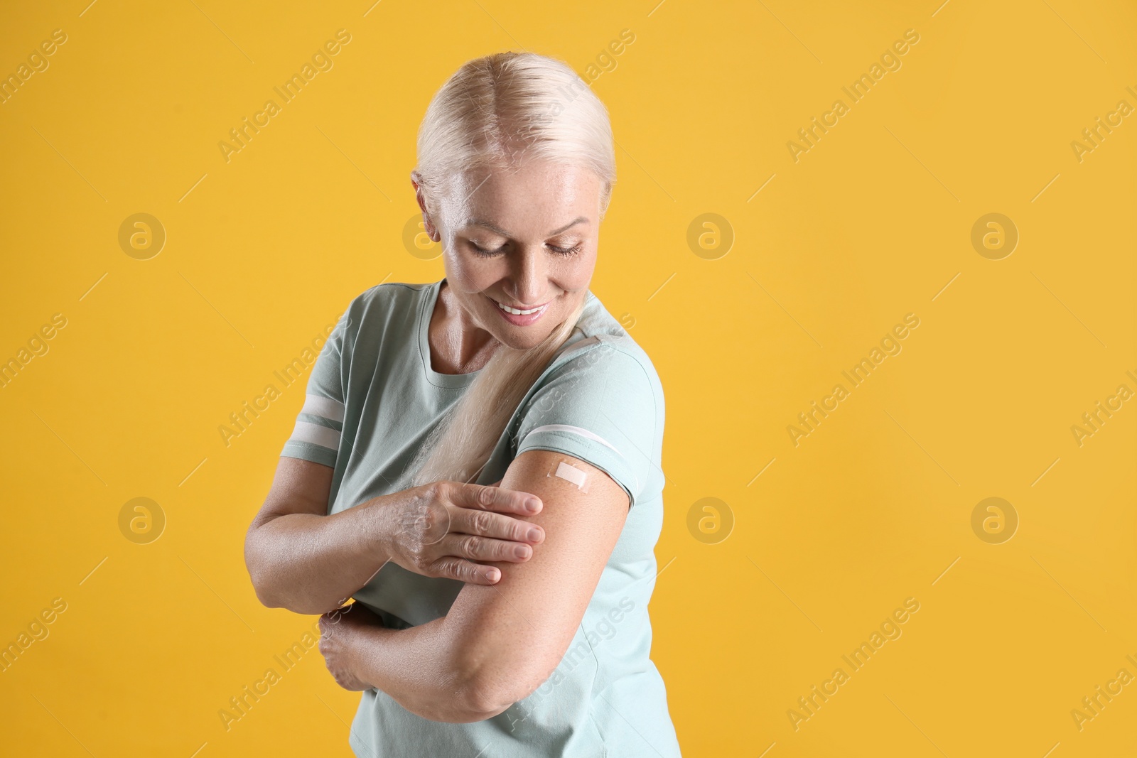 Photo of Happy mature woman showing arm with bandage after vaccination on yellow background