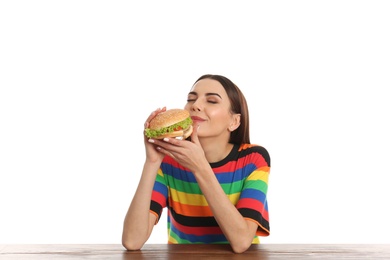 Young woman eating tasty burger at table on white background