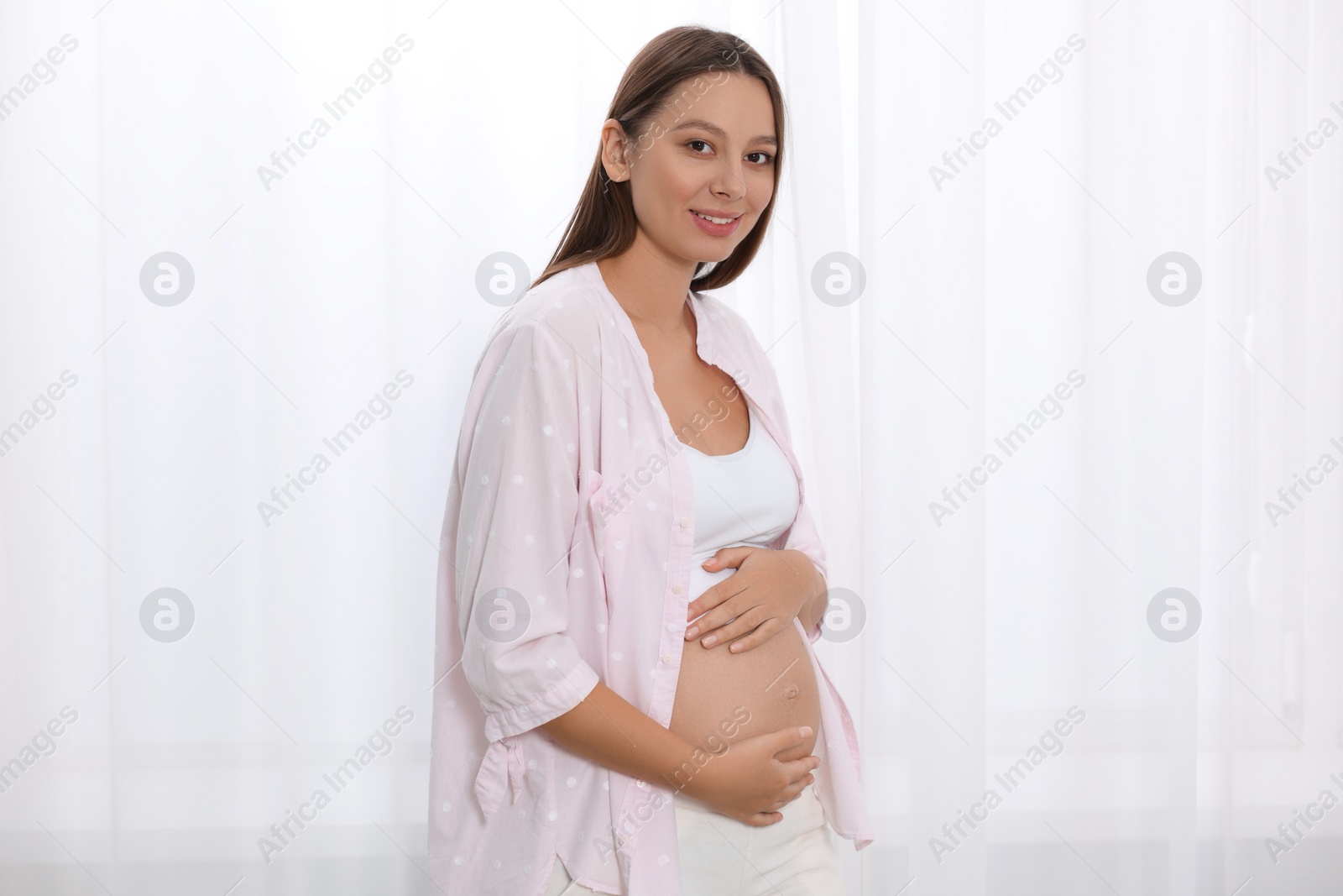 Photo of Beautiful pregnant woman in pink shirt near window indoors