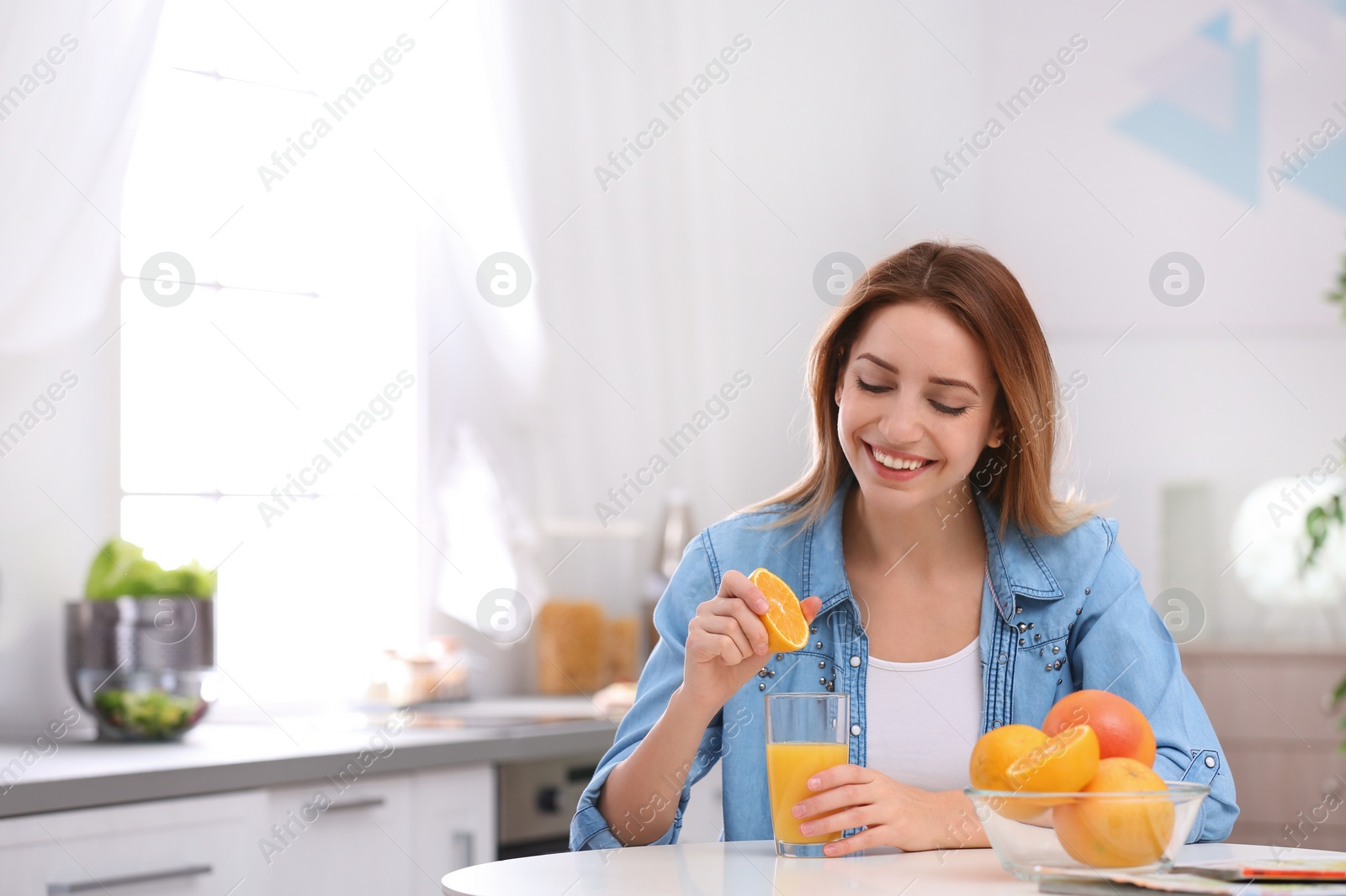 Photo of Happy young woman squeezing orange juice into glass at table in kitchen, space for text. Healthy diet