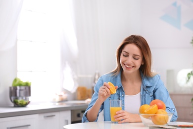 Photo of Happy young woman squeezing orange juice into glass at table in kitchen, space for text. Healthy diet