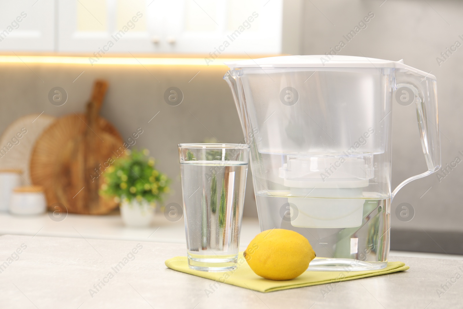 Photo of Water filter jug, glass and lemon on light grey table in kitchen, space for text