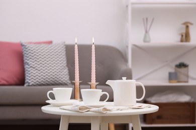 Cups of tea, teapot and burning candles on white coffee table indoors