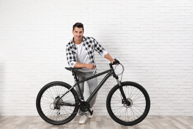 Photo of Handsome young man with modern bicycle near white brick wall indoors