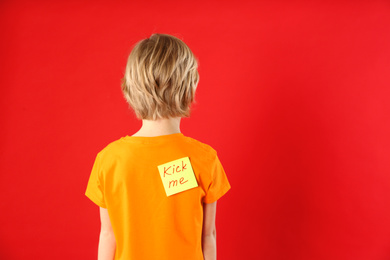 Photo of Little boy with KICK ME sticker on back against red background, space for text. April fool's day