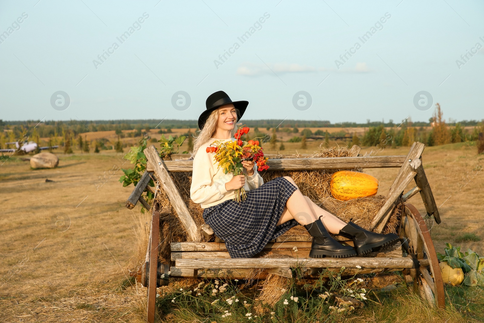 Photo of Beautiful woman with bouquet sitting on wooden cart with pumpkin and hay in field. Autumn season