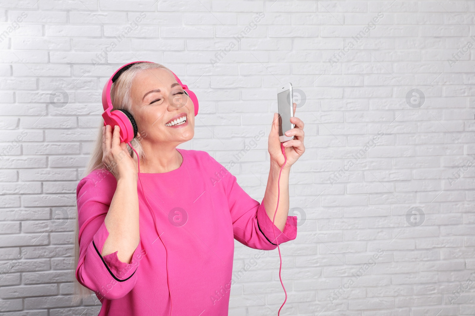 Photo of Mature woman in headphones with mobile device against brick wall. Space for text