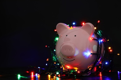 Piggy bank with Christmas lights on wooden table against black background. Space for text