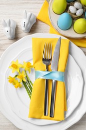 Photo of Festive table setting with painted eggs and cutlery on white wooden background, flat lay. Easter celebration