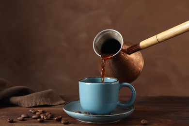 Turkish coffee. Pouring brewed beverage from cezve into cup at wooden table against brown background, space for text