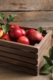 Photo of Ripe red apples with water drops in crate and green leaves on wooden table