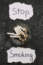 Photo of Ashtray with burnt cigarettes and words Stop Smoking written on paper on black textured table, flat lay