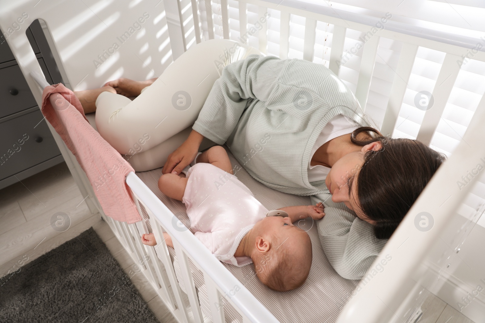 Photo of Tired young mother sleeping in crib with her baby at home, above view