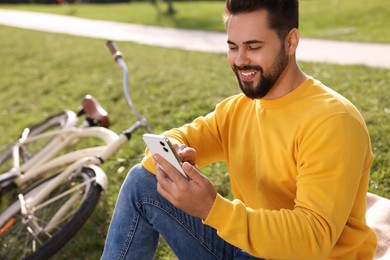 Young man sitting on green grass and using smartphone near bicycle outdoors, space for text