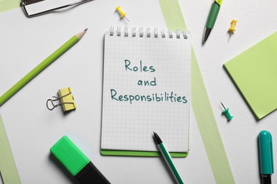 Notebook with phrase Roles And Responsibilities among office supplies on white table, flat lay