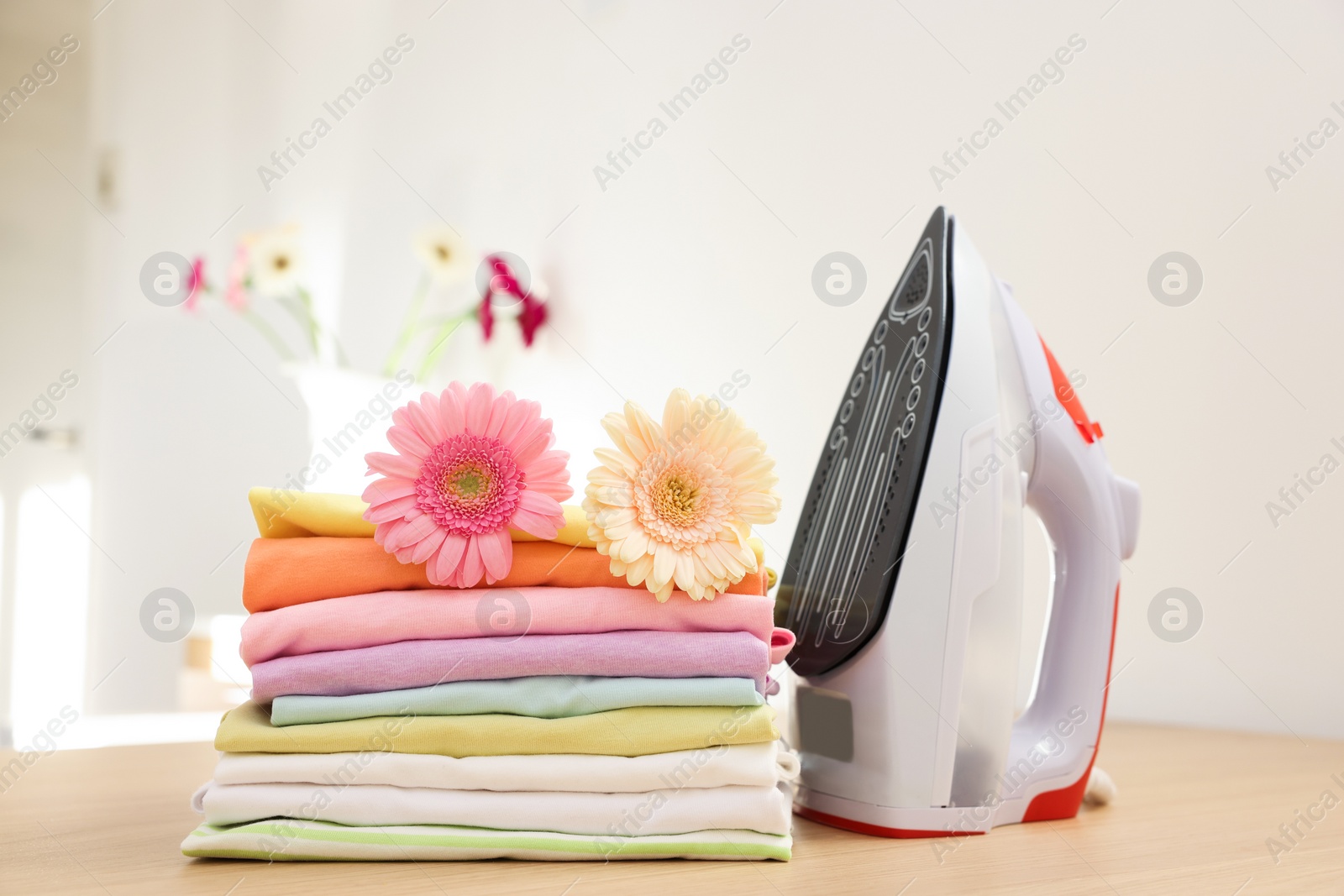 Photo of Stack of clean clothes, modern iron and flowers on wooden table