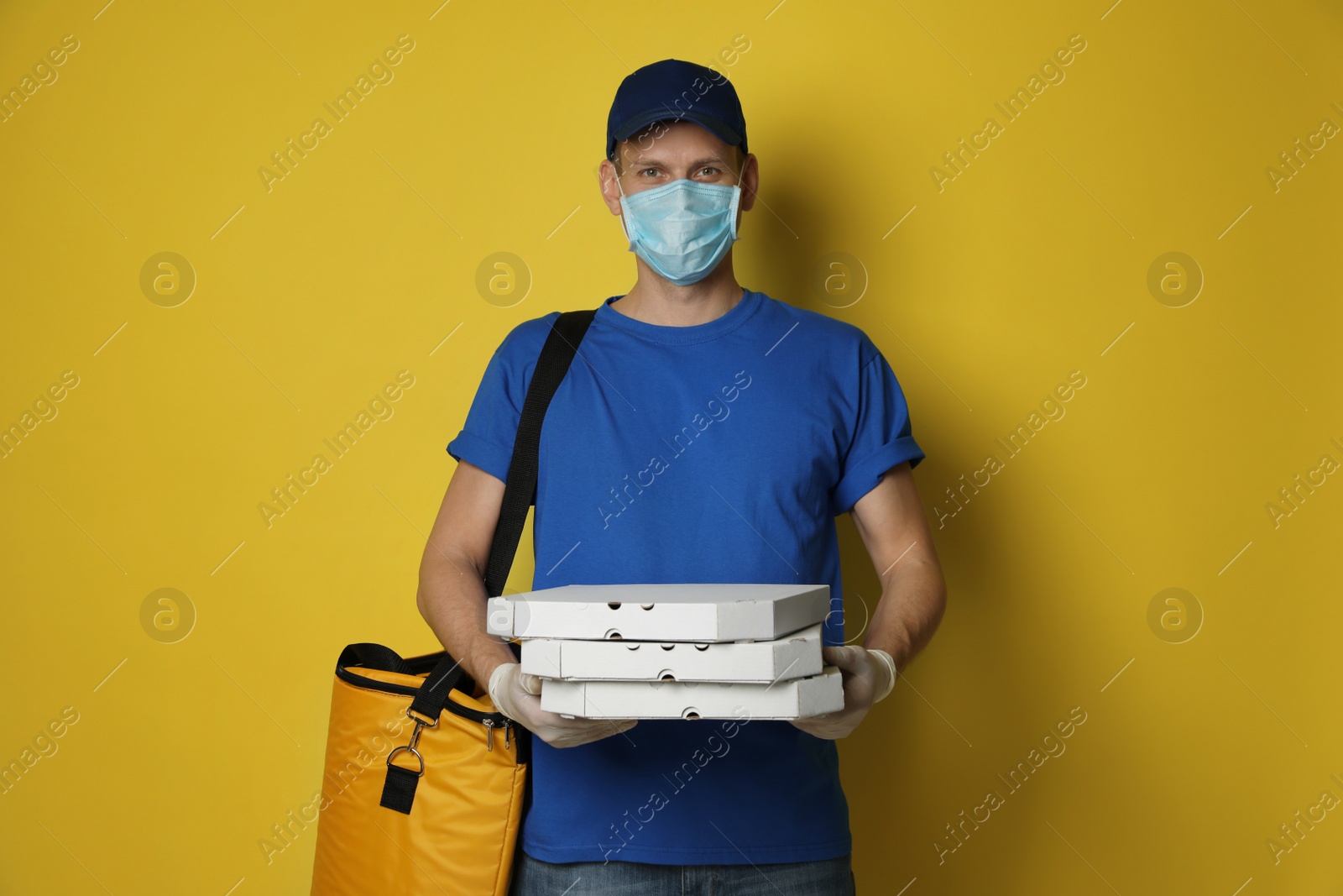 Photo of Courier in protective mask and gloves holding pizza boxes on yellow background. Food delivery service during coronavirus quarantine