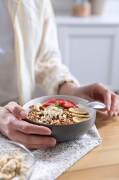 Photo of Woman eating tasty granola with banana, cashew and strawberries at wooden table indoors, closeup