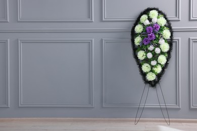 Funeral wreath of plastic flowers near light grey wall indoors, space for text