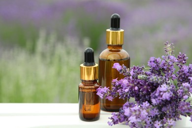 Photo of Bottles of essential oil and lavender flowers on white wooden table in field, space for text