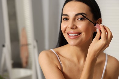 Beautiful young woman applying mascara indoors, closeup. Space for text