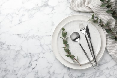 Stylish setting with cutlery, napkin, eucalyptus branches and plates on white marble table, top view. Space for text