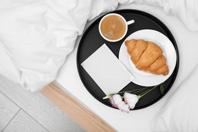 Tray with tasty croissant, cup of coffee and flowers on white bed, top view