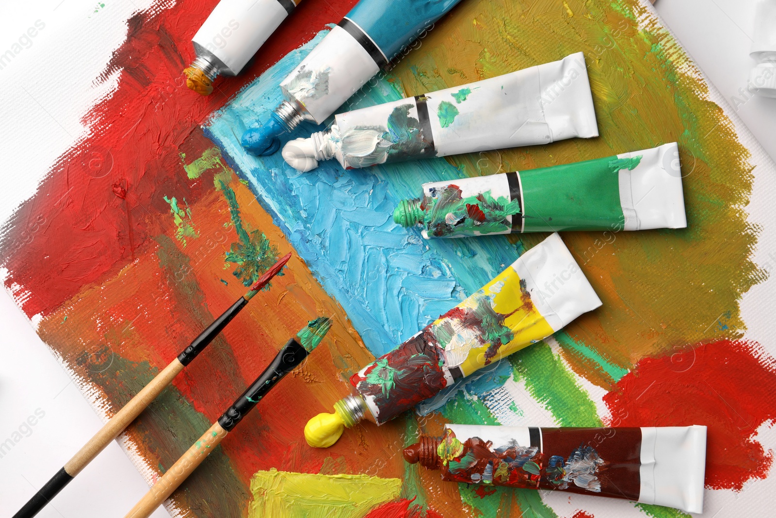 Photo of Tubes of colorful oil paints and brushes on canvas with abstract painting, flat lay