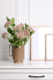 Photo of Stylish ceramic vase with beautiful flowers and blank frames on chest of drawers near white wall