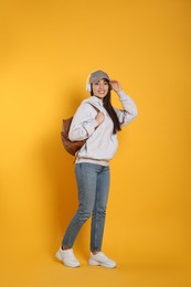 Beautiful young woman with stylish leather backpack and headphones on yellow background