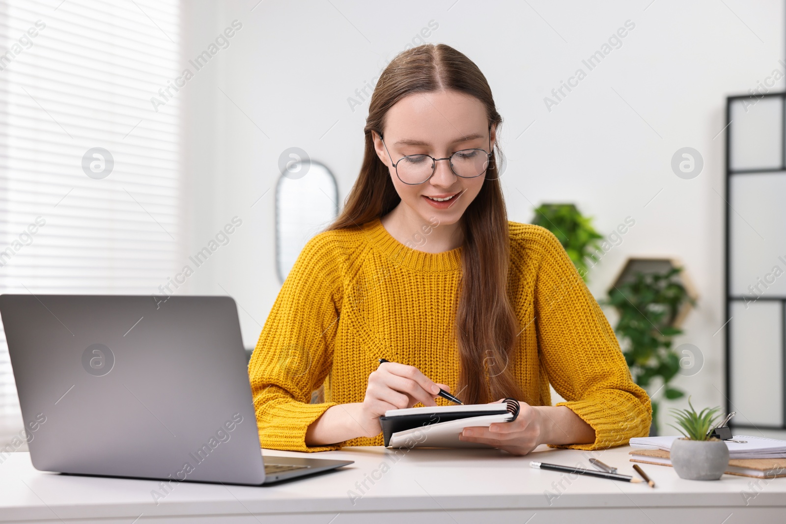 Photo of E-learning. Young woman taking notes during online lesson at white table indoors