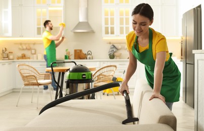 Photo of Professional janitor in uniform vacuuming sofa indoors