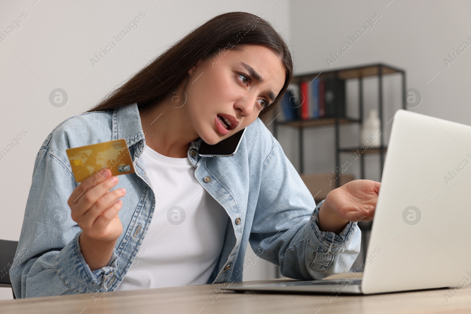 Photo of Confused woman with credit card talking on phone and using laptop at table indoors. Be careful - fraud